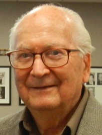 Holocaust survivor Al Beder wearing glasses and a collared shirt, photographed in front of a wall of framed black-and-white photographs.