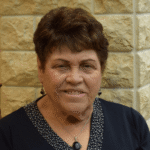 Holocaust survivor Edie Pump, elderly woman with warm smile and short styled brown hair, wearing navy blue top with white polka dot collar trim, black drop earrings and matching pendant necklace, photographed against textured stone wall background