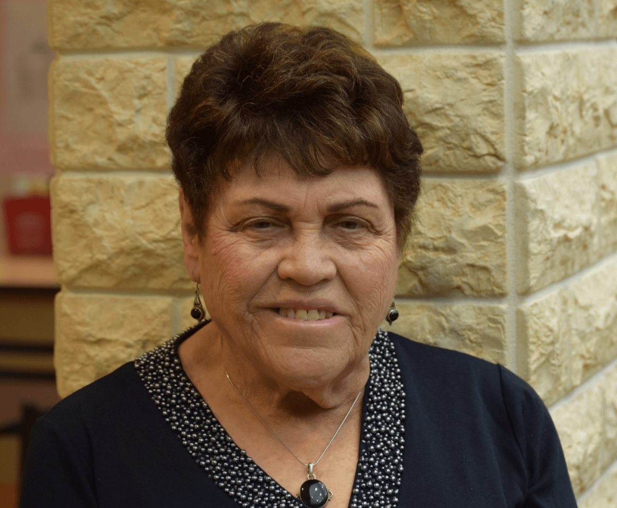Holocaust survivor Edie Pump, elderly woman with warm smile and short styled brown hair, wearing navy blue top with white polka dot collar trim, black drop earrings and matching pendant necklace, photographed against textured stone wall background