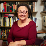 Holocaust survivor Eva Zaret, elderly woman with warm smile, wearing glasses and burgundy long-sleeved top, photographed in a library or study with full bookshelves in the background