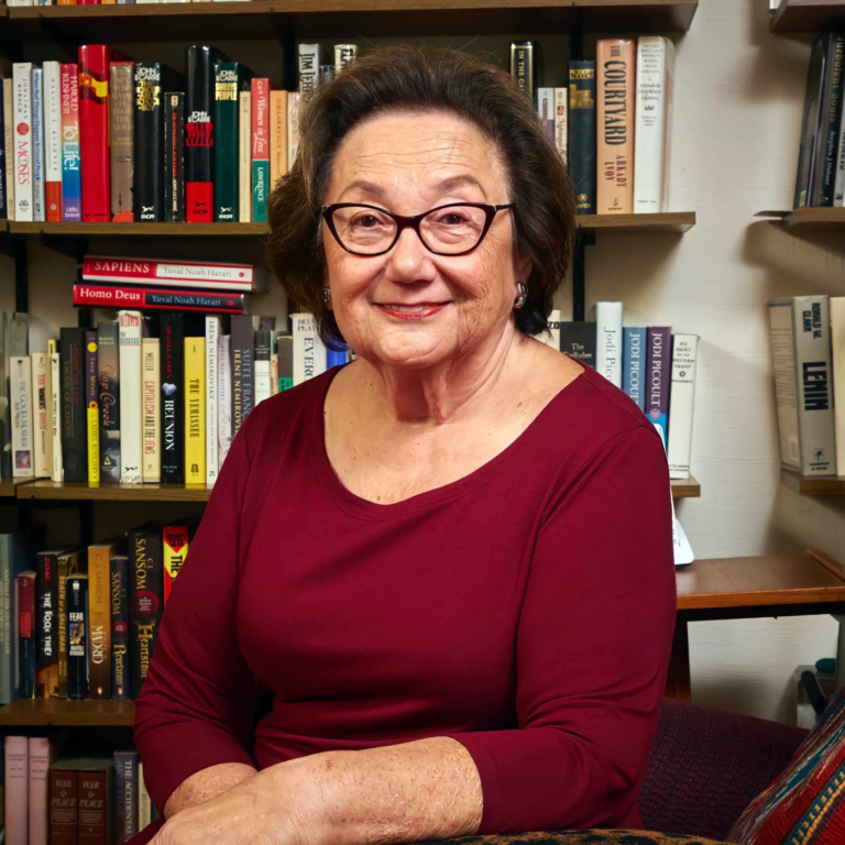Holocaust survivor Eva Zaret, elderly woman with warm smile, wearing glasses and burgundy long-sleeved top, photographed in a library or study with full bookshelves in the background