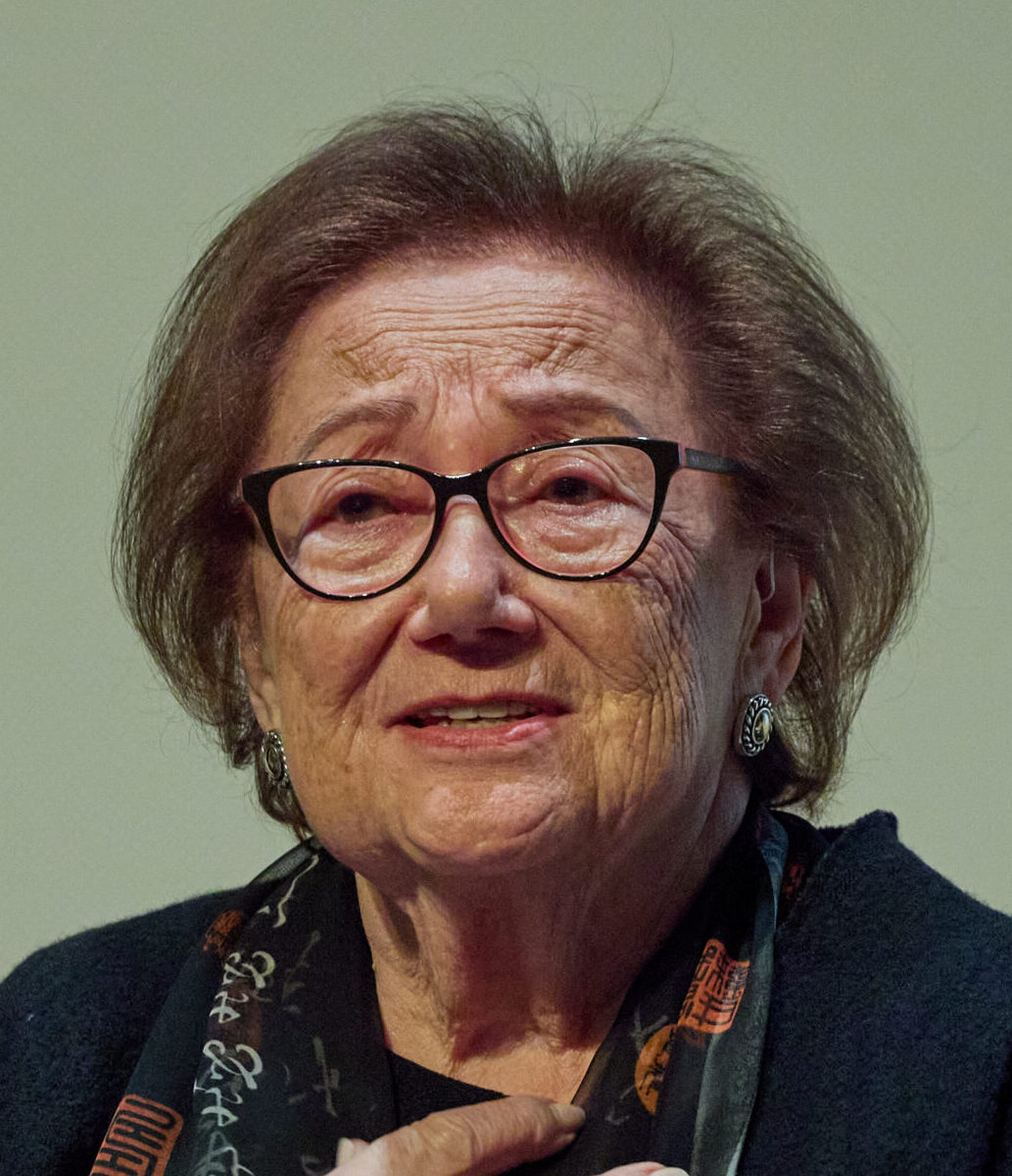 Holocaust survivor Eva Zaret seated and speaking, her hand resting on her chest, wearing a black blazer and patterned scarf and glasses, photographed against a neutral background.