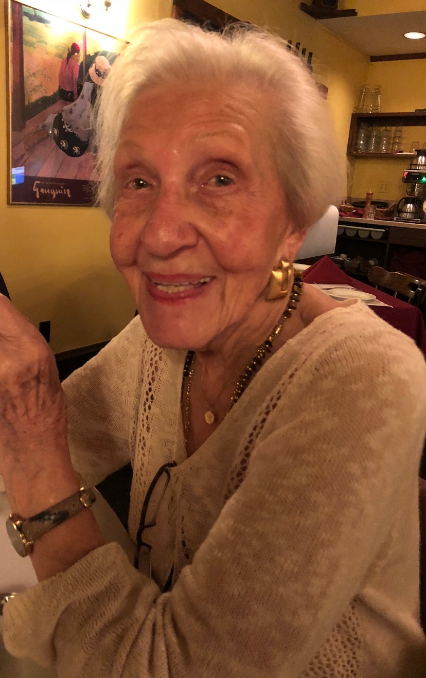 Holocaust survivor Jean Yuspeh smiling warmly at the camera, wearing a cream sweater and gold jewelry, photographed in a restaurant setting.