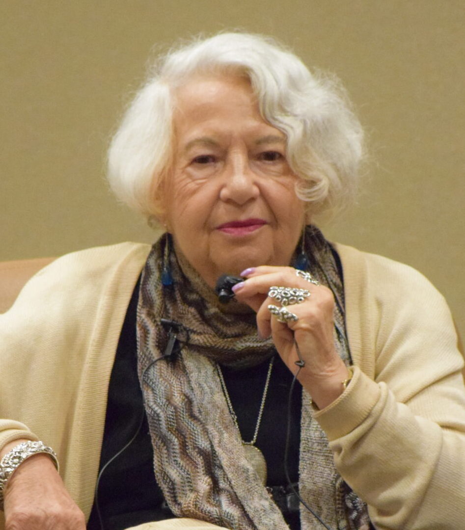 Holocaust survivor Kaja Finkler seated in a leather armchair, wearing a cream cardigan, black outfit, and fringed scarf, with her hand raised thoughtfully near her chin, photographed at an indoor event venue.