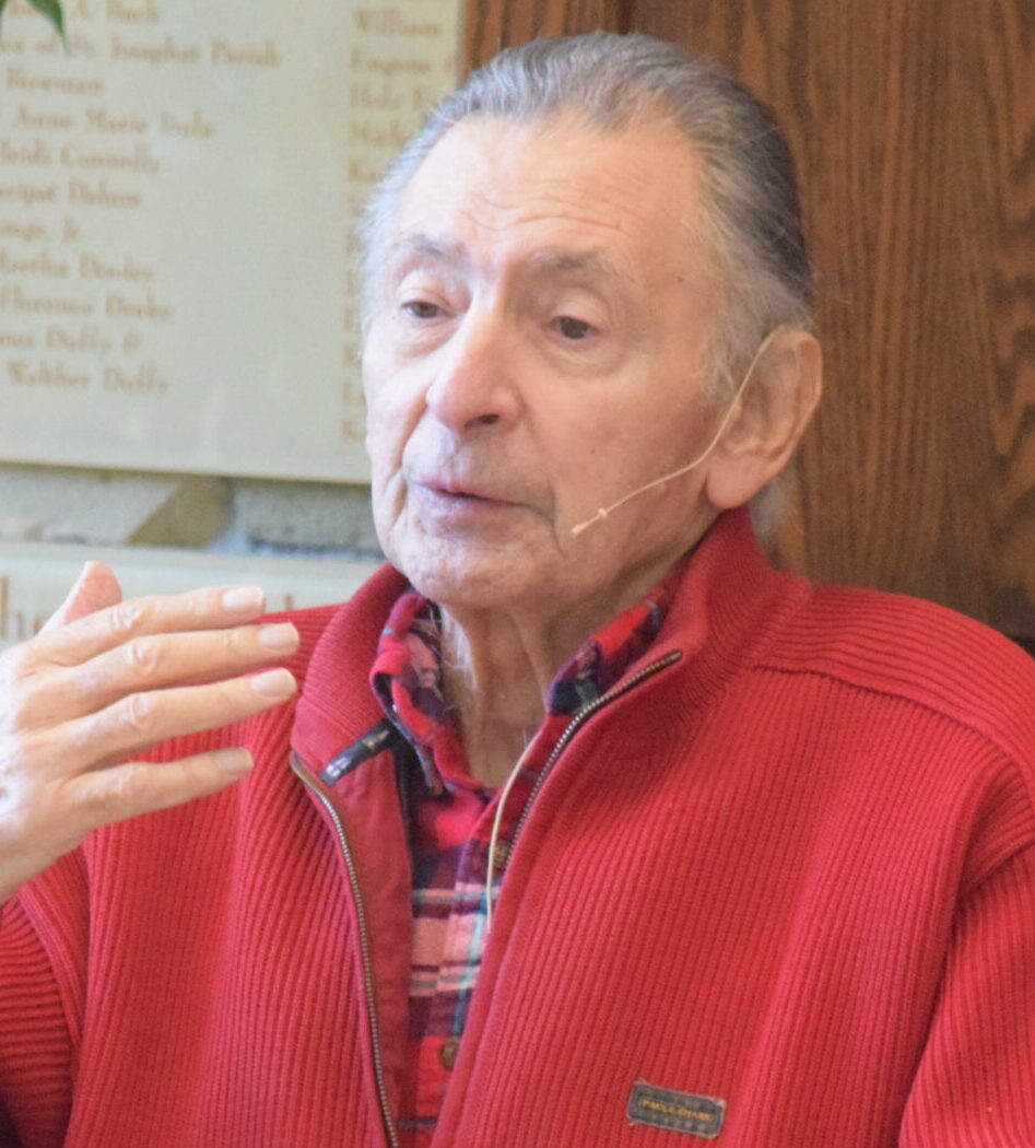 Holocaust survivor Lee Marnett seated and speaking, gesturing with one hand while wearing a red zip-up sweatshirt and a lapel microphone, photographed at an indoor event venue.