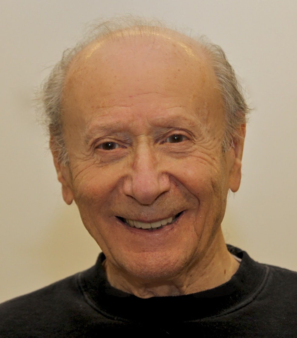 Holocaust survivor Louis Koplin smiling broadly, wearing a black crew-neck shirt, in a close-up portrait against a neutral background.
