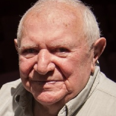 Nathan Taffel, a Holocaust survivor, gazes steadily at the camera. He has short white hair and wears a beige collared shirt.