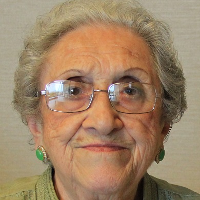 Raye David, a Holocaust survivor, looks quietly at the camera wearing wire-rimmed glasses and a soft green jacket.