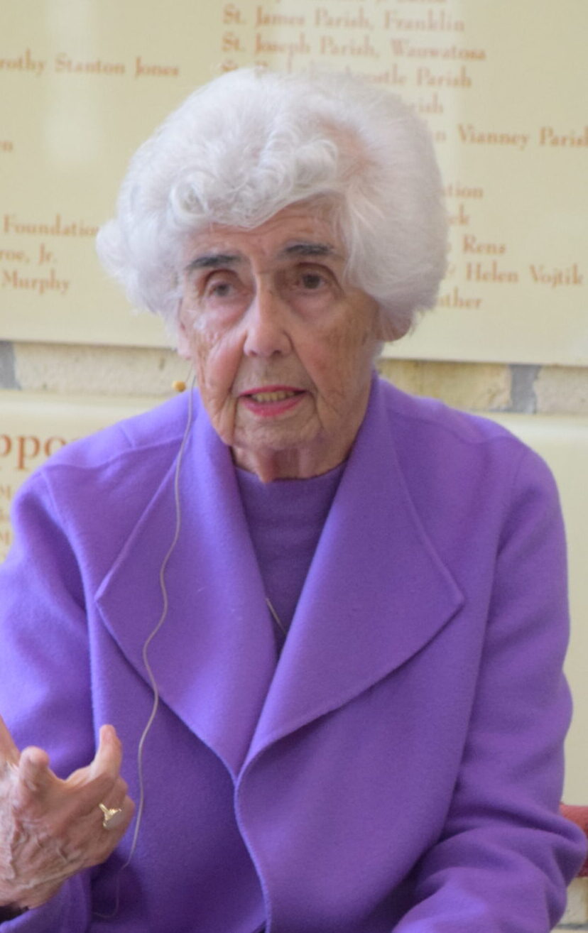 Holocaust survivor Renata Laxova seated and speaking, gesturing with one hand while wearing a purple jacket and lapel microphone, photographed in front of a brick wall and donor recognition plaques at an indoor venue.