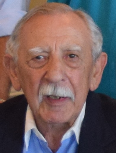 Sam Gingold, a Holocaust survivor, appears mid-expression wearing a light blue collared shirt and a white mustache.