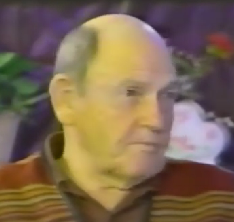 Anton Bild, a Holocaust survivor, speaks during a recorded interview. He is bald and wears a striped sweater in warm earth tones.