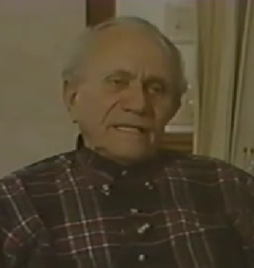 Mikhail Becker, a Holocaust survivor, speaks during a recorded interview. He has thinning gray hair and wears a dark plaid flannel shirt.