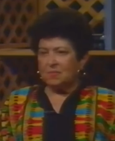 Rose Chrustowski, a Holocaust survivor, speaks during a recorded interview. She has short dark hair and wears a black top with a boldly striped multicolored jacket.