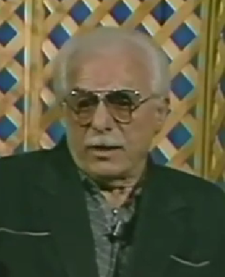 Walter Peltz, a Holocaust survivor, speaks during a recorded interview. He is bald with a light mustache and wears tinted aviator-style glasses and a dark suit jacket.