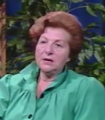 Franka Neufeld, a Holocaust survivor, speaks during a recorded interview. She has short auburn hair and wears a green button-down blouse.