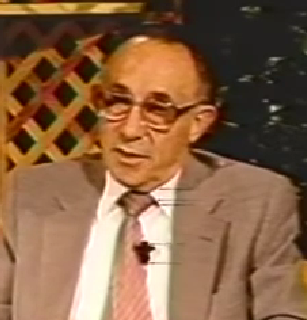 Jack Marcus, a Holocaust survivor, speaks during a recorded interview wearing dark-rimmed glasses and a brown suit jacket.