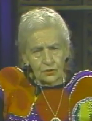 Sarah Federman, a Holocaust survivor, speaks during a recorded interview. She has short white hair and wears a colorful patterned top with a gold pendant necklace.