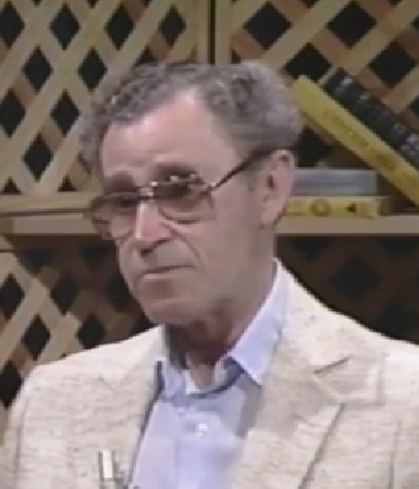 Mike Kleiner, a Holocaust survivor, speaks during a recorded interview. He has short gray hair, wire-rimmed glasses, and wears a light tan blazer over a blue collared shirt.