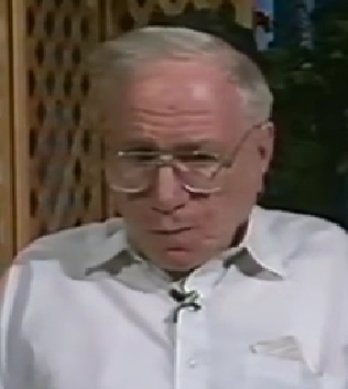 Tzvi Deutsch, a Holocaust survivor, speaks during a recorded interview. He has short gray hair, wire-rimmed glasses, and wears a white button-down shirt.