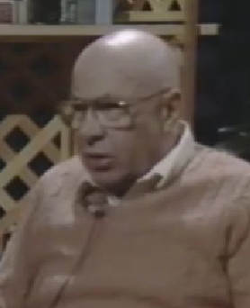 Dudek Birnbaum, a Holocaust survivor, speaks during a recorded interview. He is bald with a light mustache, wears wire-rimmed glasses, and a tan sweater over a collared shirt.
