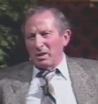 Pincus Weinstock, a Holocaust survivor, speaks during a recorded interview. He has short gray hair and wears a gray suit jacket with a striped tie.