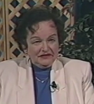 Helga Heymann, a Holocaust survivor, speaks during a recorded interview. She has short dark hair and wears a white blazer over a blue top with a delicate necklace.