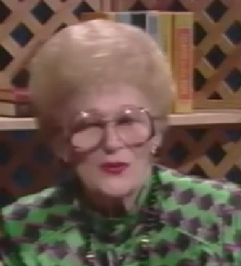 Bernice Birnbaum, a Holocaust survivor, speaks during a recorded interview. She has short white hair, large tinted glasses, and wears a green patterned top.