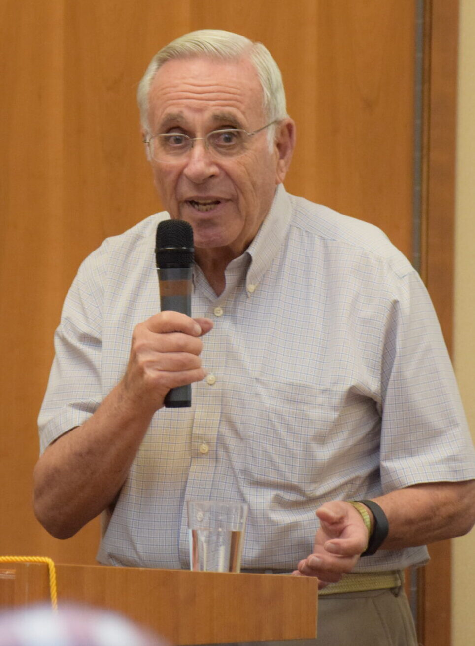Holocaust survivor Steen Metz standing at a podium and speaking into a handheld microphone, wearing a light blue short-sleeve button-down shirt, photographed in an indoor event space.