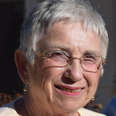 Susie Fono, a Holocaust survivor, smiles warmly at the camera wearing wire-rimmed glasses and a light cream top.