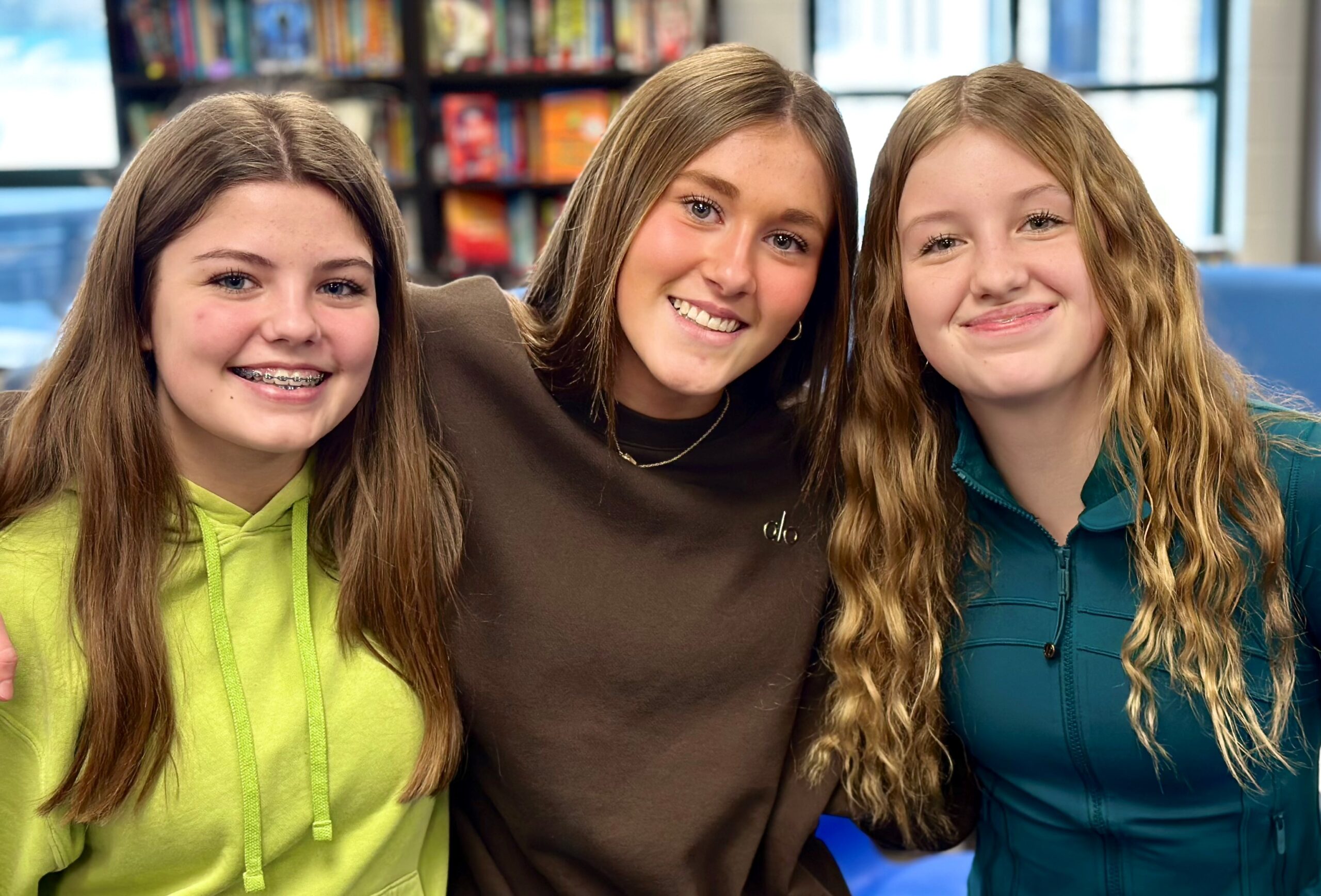 Three smiling young women stand together with their arms around each other in what appears to be a classroom or educational space. The woman on the left wears a bright yellow-green hoodie, the woman in the center wears a brown turtleneck sweater, and the woman on the right wears a teal quarter-zip pullover. Colorful educational materials are visible on the wall in the background.