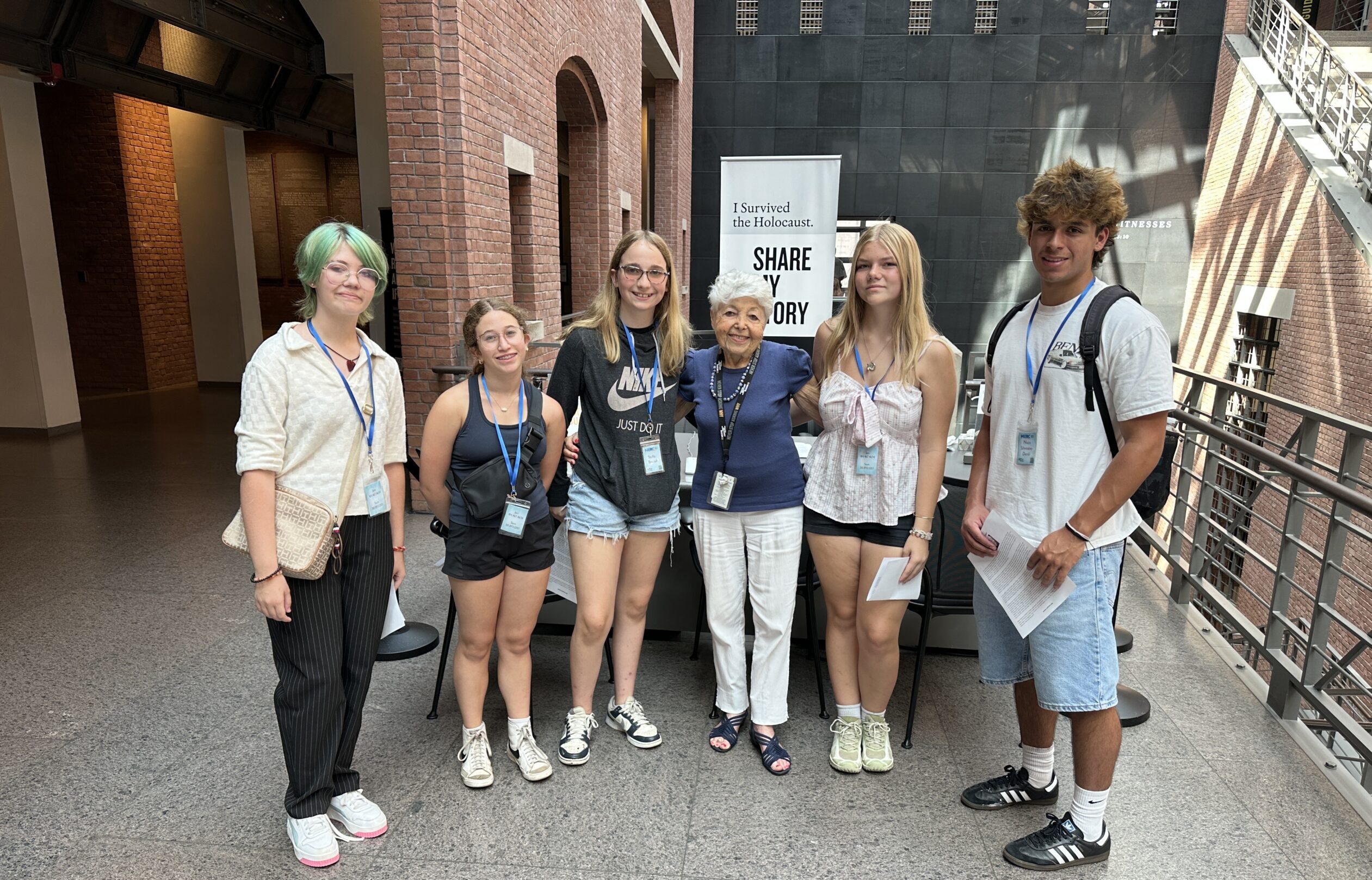 Six Writing and Art Contest winners pose together at the Holocaust Museum, wearing blue visitor lanyards, with a 'Share My Story' banner visible behind them.
