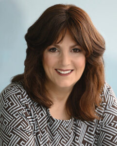 Professional headshot of Liza Wiemer the author of The Assignment, smiling woman with shoulder-length auburn hair and bangs, wearing a black and white geometric patterned top against a light blue background