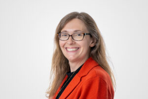 Samantha Abramson, HERC's Executive Director, smiles at the camera against a light gray background. She has shoulder-length brown hair, wears glasses, and is dressed in a bright orange blazer over a black top.