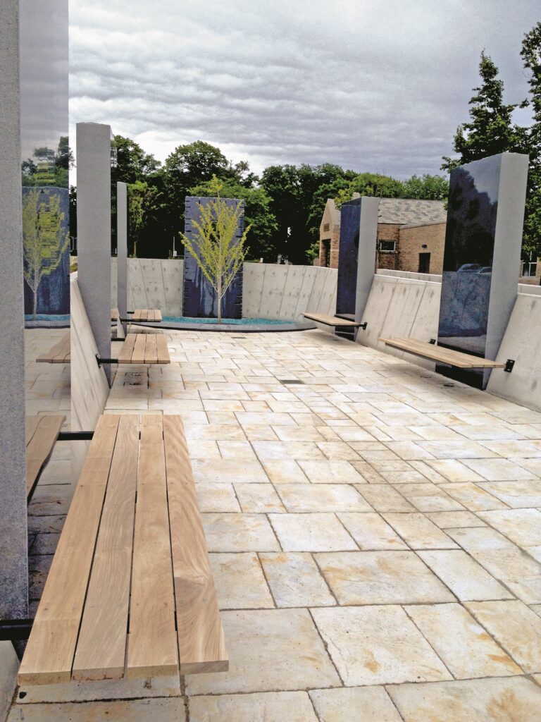 Pinat Hatikvah (Corner of Hope), an outdoor memorial space featuring light-colored stone pavers and wooden benches attached to concrete pillars. Tall concrete panels display images of trees, and a young tree with bright green-yellow leaves stands at the center of the space, surrounded by blue-green decorative stones. The memorial is set against a backdrop of mature trees and an overcast sky, with a brick building visible in the background.