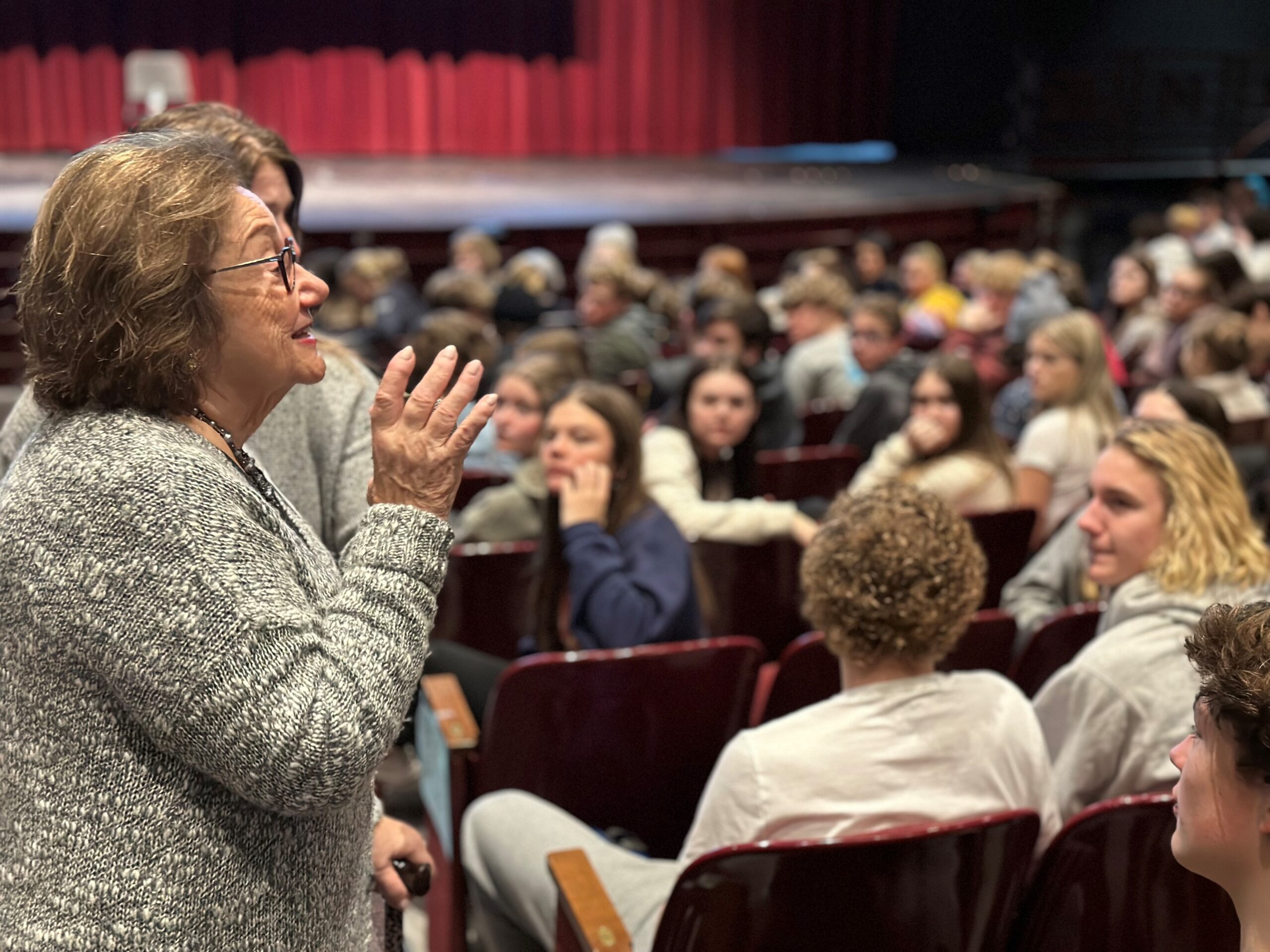 A woman, Eva Zaret Holocaust survivor, with brown hair and glasses wearing a gray textured cardigan gestures expressively while speaking to a large audience of students seated in an auditorium with red curtains visible in the background.