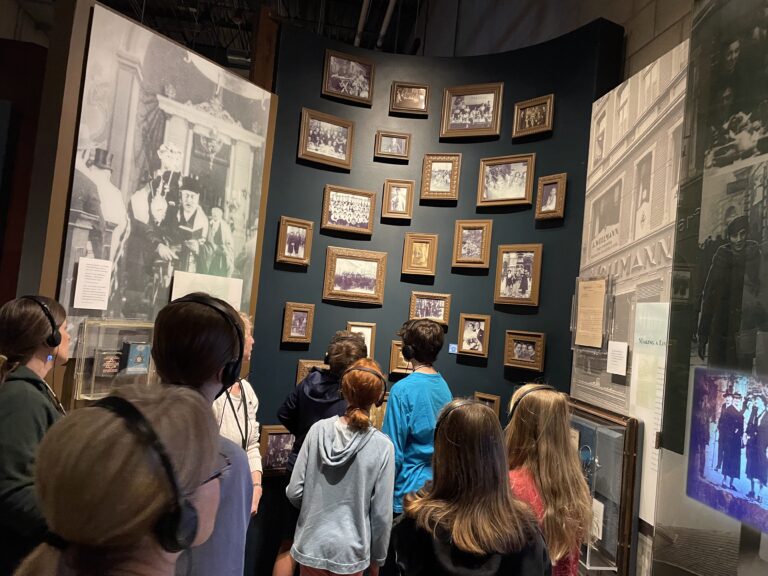 A group of approximately eight students wearing audio headsets stand together viewing an exhibit at the Illinois Holocaust Museum & Education Center. They face a curved dark blue wall displaying numerous gold-framed black-and-white historical photographs arranged in a gallery style. Large historical photographs of European Jewish life are visible on surrounding panels, including images of storefronts and community gatherings. A small display case with artifacts is visible to the left.