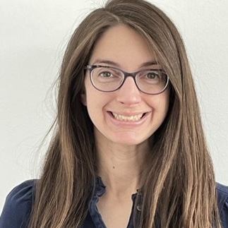 Professional headshot of a woman, Margo Berger with long brown hair and glasses, smiling warmly at the camera. She wears a dark navy top with a zipper detail, photographed against a light neutral background.