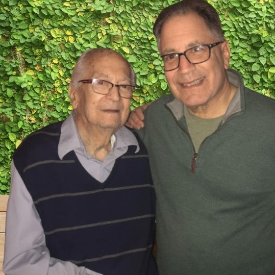 Two men standing together with arms around each other's shoulders, smiling at the camera against a green foliage wall. The man on the left, Al Beder, wears glasses and a navy striped sweater vest over a light blue shirt. The man on the right, Al's son Jeff, wears glasses and a green quarter-zip pullover.