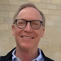 Professional headshot of a man, Andy Palec, with gray hair wearing dark-framed glasses and a navy blazer over a light blue collared shirt, smiling at the camera against a light brick wall background.
