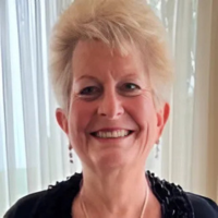 Professional headshot of a woman, Dena Rubnitz, with short blonde hair and dangling earrings, smiling warmly at the camera. She wears a black top with decorative detail at the neckline, photographed against a light background with soft window lighting.
