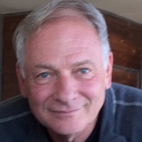 Professional headshot of a man, Marty Thau, with gray hair wearing a dark striped polo shirt, smiling warmly at the camera against a soft-focus indoor background.