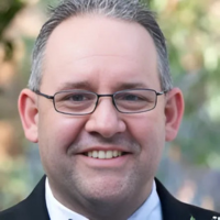 Professional headshot of a man, Brad Tenin, with short gray hair wearing dark-framed glasses and a dark suit with white shirt and tie, smiling at the camera against a soft-focus outdoor background.