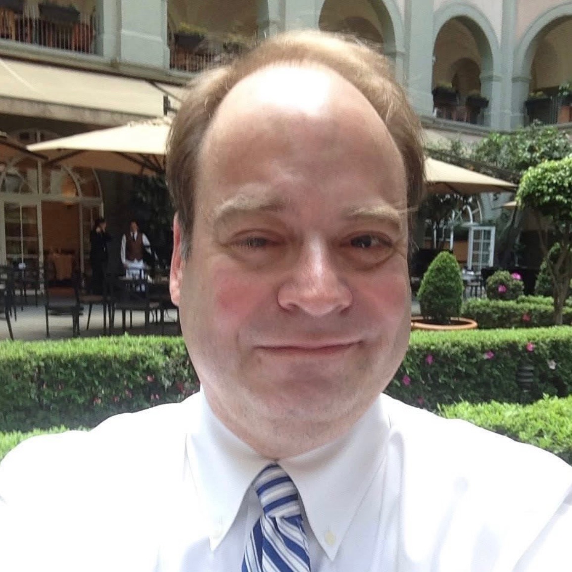 Professional photo of a man, Steven Jacobs, with light brown hair wearing a white dress shirt and blue striped tie, smiling at the camera. Photographed outdoors with a courtyard setting featuring green landscaping, umbrellas, and white architectural elements in the background.