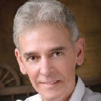 Professional headshot of a man with silver-gray hair wearing a dark blazer and light blue shirt, photographed against a warm-toned background.