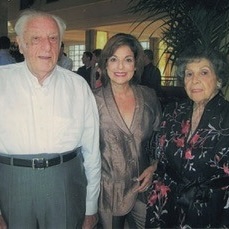 Three people standing together indoors: a man in a white shirt on the left, a woman, Susie Schneiderman, in a brown cardigan in the center, and a woman in a dark floral-patterned top on the right, all smiling at the camera with greenery visible in the background.