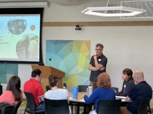 A man wearing glasses and a dark short-sleeved shirt stands presenting to a small group seated around a conference table in a modern meeting room. A projection screen displays historical images, and a colorful geometric wall art piece in blue, green, and yellow tones is visible behind the speaker.