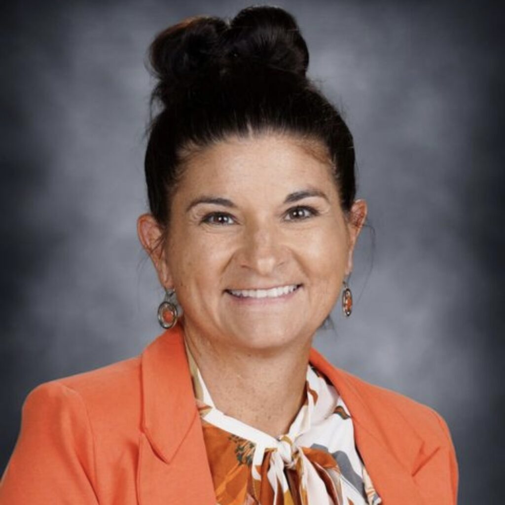 Jess Donnerbauer, HERC Fellow and Wisconsin educator, smiling in a professional headshot. She has dark hair styled in a top bun and wears an orange blazer over a cream blouse with an orange, brown, and gray abstract print tied at the neck. She has circular drop earrings with orange centers, photographed against a mottled gray-blue background.