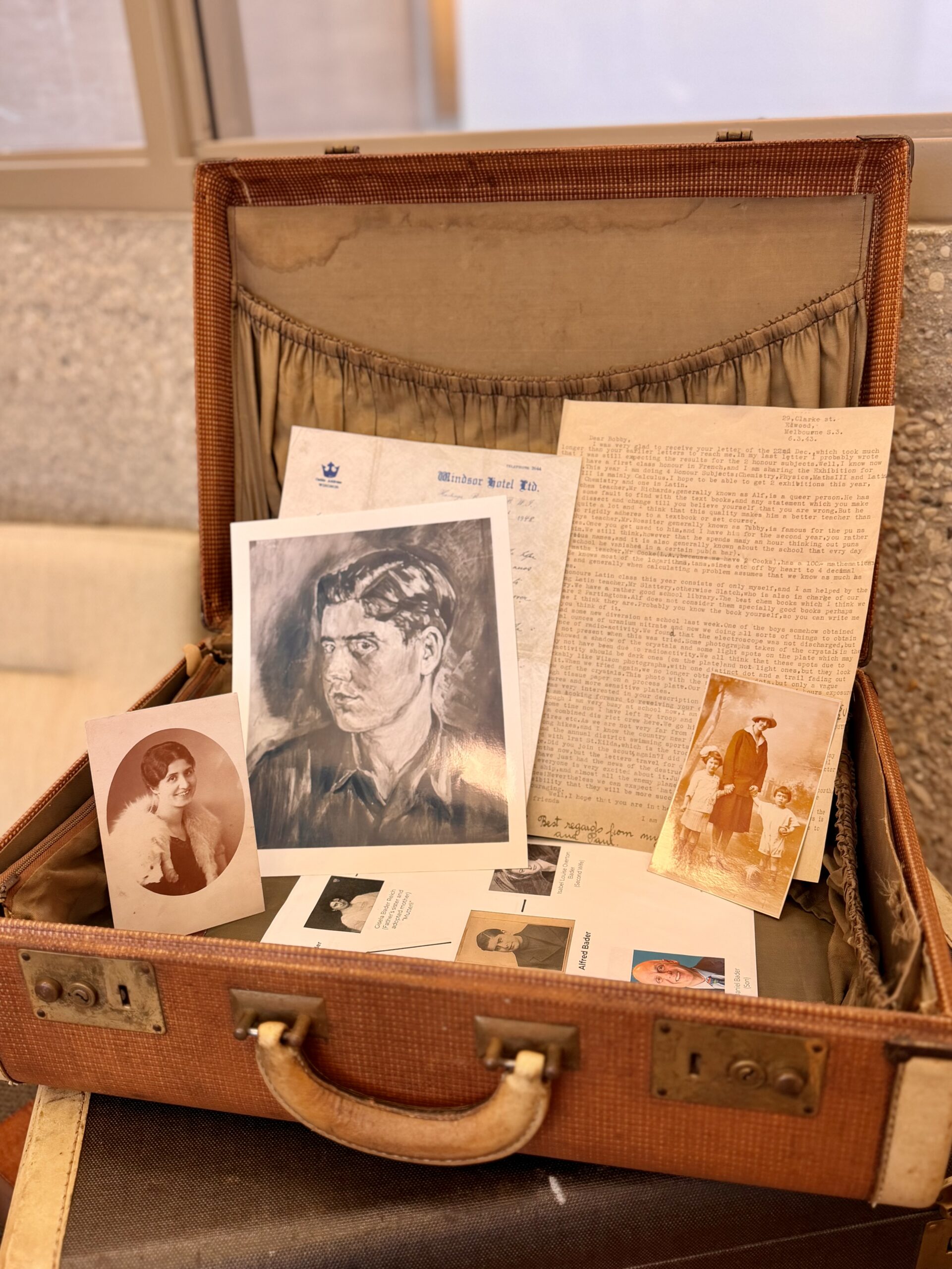 An open vintage suitcase displaying historical artifacts from Holocaust survivor Alfred Bader, including family photographs, a portrait, and personal letters.