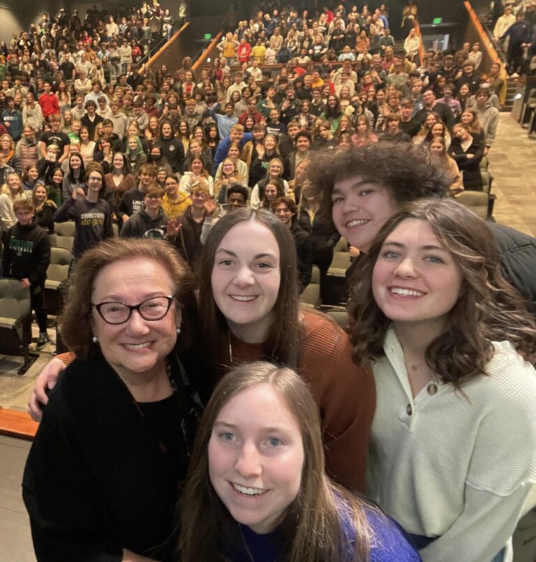 Four smiling women, including Holocaust survivor and Speakers Bureau member, Eva Zaret, take a selfie in an auditorium filled with attendees. The photo is taken from their perspective looking back at rows of seated audience members. The women are positioned in the foreground, with one wearing glasses and a black top, another in an orange/rust-colored top, one in a light mint green sweater, and one in a purple top in the front center. The large crowd behind them fills the theater-style seating.
