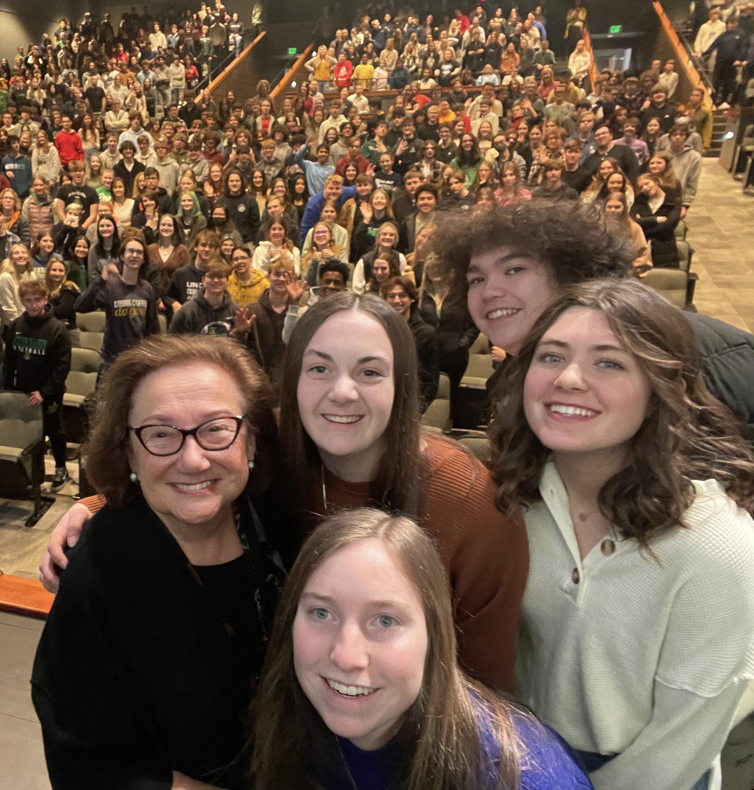 Four smiling women, including Holocaust survivor and Speakers Bureau member, Eva Zaret, take a selfie in an auditorium filled with attendees. The photo is taken from their perspective looking back at rows of seated audience members. The women are positioned in the foreground, with one wearing glasses and a black top, another in an orange/rust-colored top, one in a light mint green sweater, and one in a purple top in the front center. The large crowd behind them fills the theater-style seating.